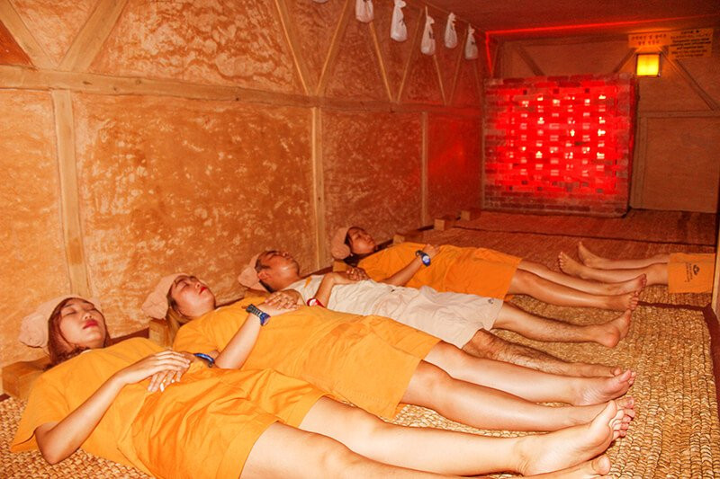 Hình ảnh những người đang nằm thư giãn trong một phòng xông hơi được ốp gỗ, ánh sáng đỏ ấm áp, mặc trang phục màu cam tại spa xông hơi quận 7.