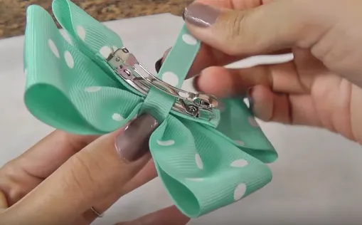 Quấn dải dây chữ nhật vòng lên qua điểm chính giữa chiếc nơ và vòng 1 vòng xuống phía trong kẹp tóc rồi quấn đến khi hết đoạn vải và gắn keo lại