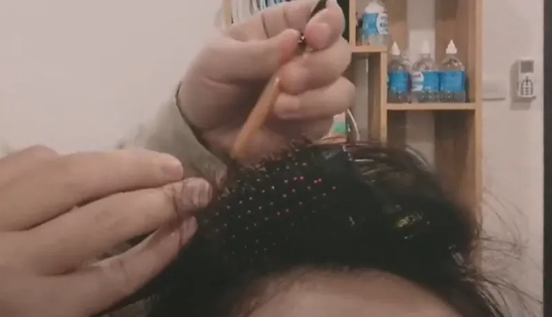 Mô tả cụ thể nội dung hình bằng tiếng Việt, một người đang dùng que mảnh để nhẹ nhàng gỡ các lớp tóc rối bị mắc vào lược tròn