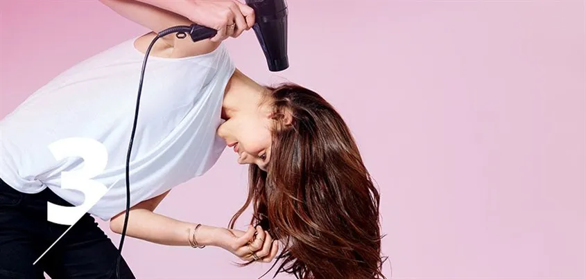 Người phụ nữ cúi đầu khi sấy tóc từ sau gáy lên phía trước, tay luồn vào tóc để tạo độ bồng